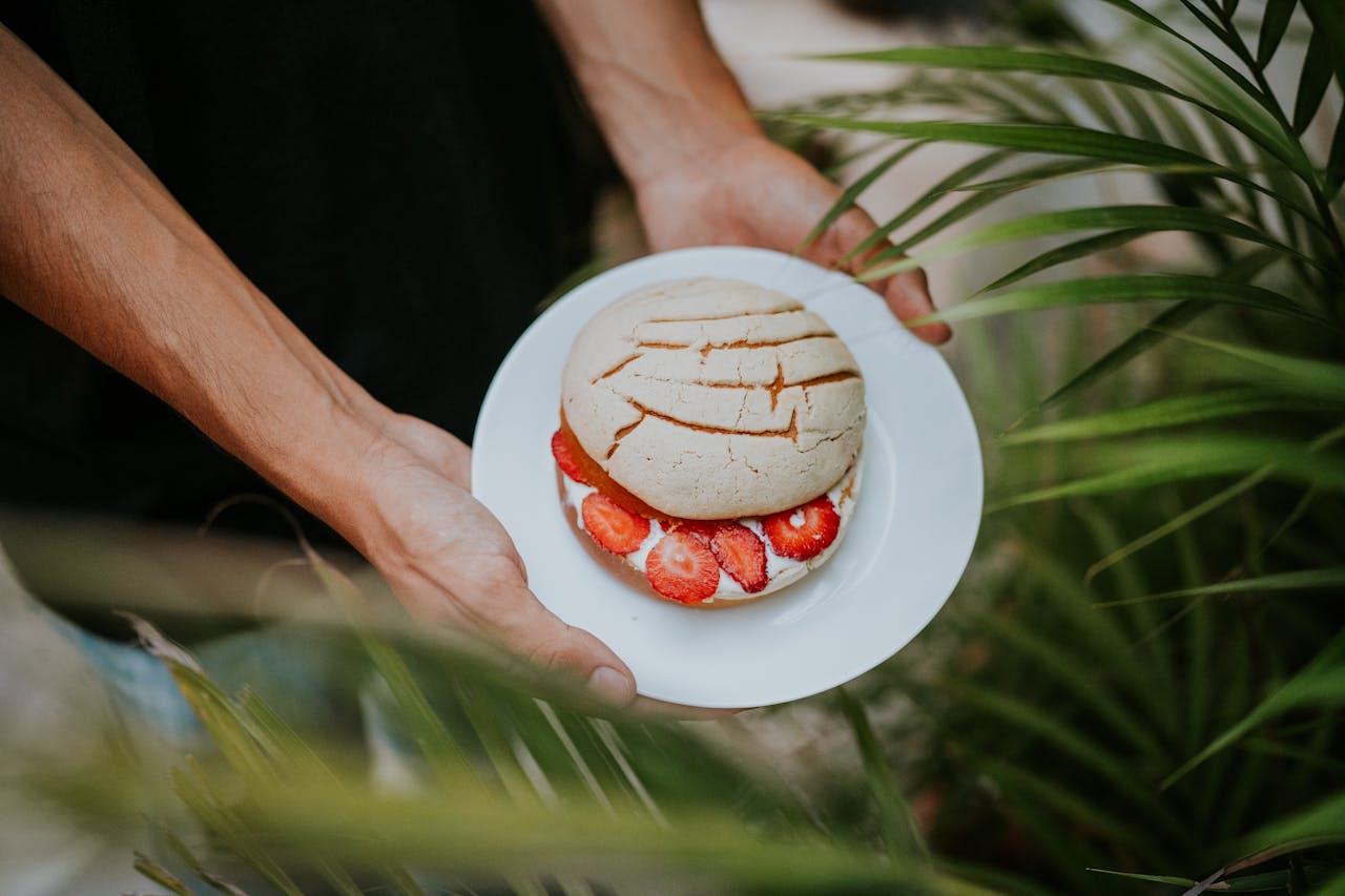 A close-up shot of Mexican pan dulce on a plate, garnished with fresh strawberries, held by hands outdoors.